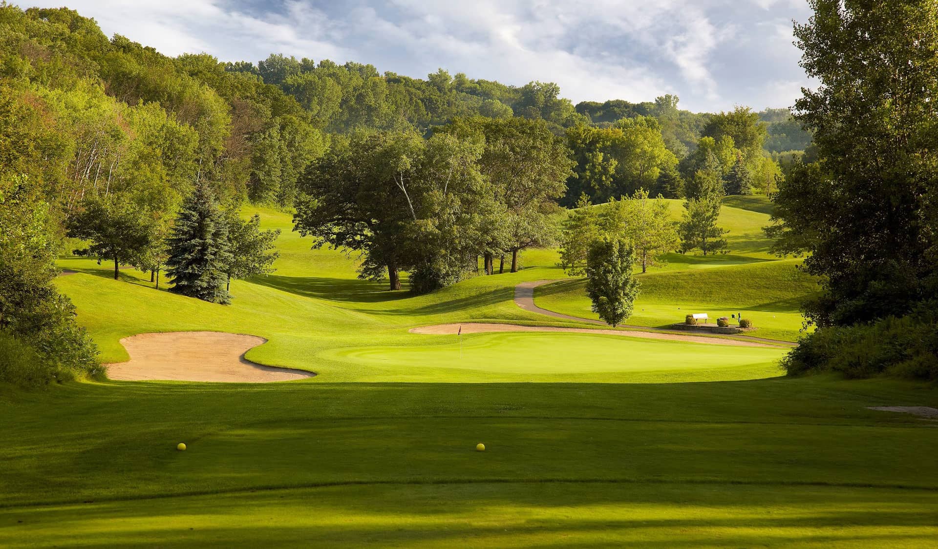 Rolling golf course fairway leads to a green with flagstick, large bunker, and trees under a partly cloudy sky.