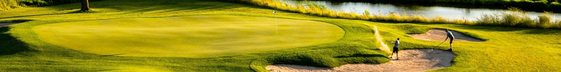 Two golfers on a sunlit green, one chipping from a sand bunker toward the flagstick, with a pond in background.