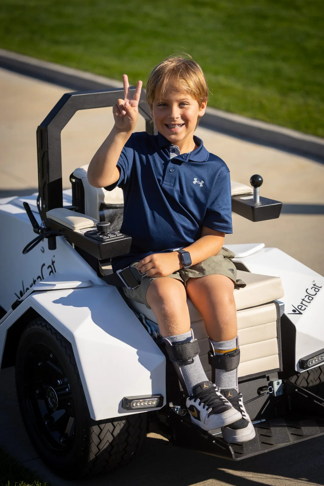 Smiling boy in an accessible VertaCat golf cart, making a peace sign, with leg braces.