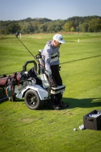 Golfer in a VertaCat stand-up golf cart on the course, holding a driver. A box of golf balls sits nearby.