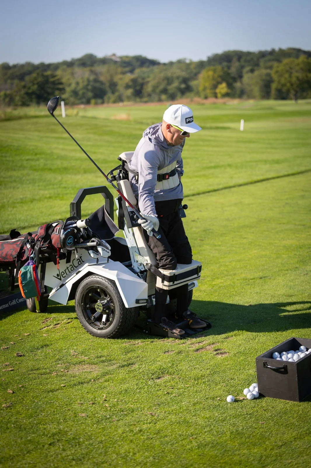 Golfer in a VertaCat stand-up golf cart on the course, holding a driver. A box of golf balls sits nearby.