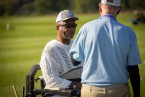 Adaptive golfer in specialized cart, wearing MGA cap and sunglasses, converses with another person on course.