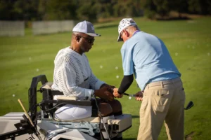 Golfer in adaptive cart receives instruction from a coach on a sunny driving range.