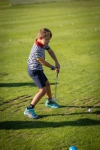 Young boy in striped shirt swinging a golf club on a golf course with a golf ball on a pink tee.
