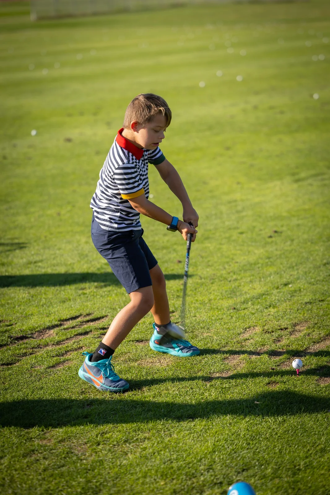 Young boy in striped shirt swinging a golf club on a golf course with a golf ball on a pink tee.