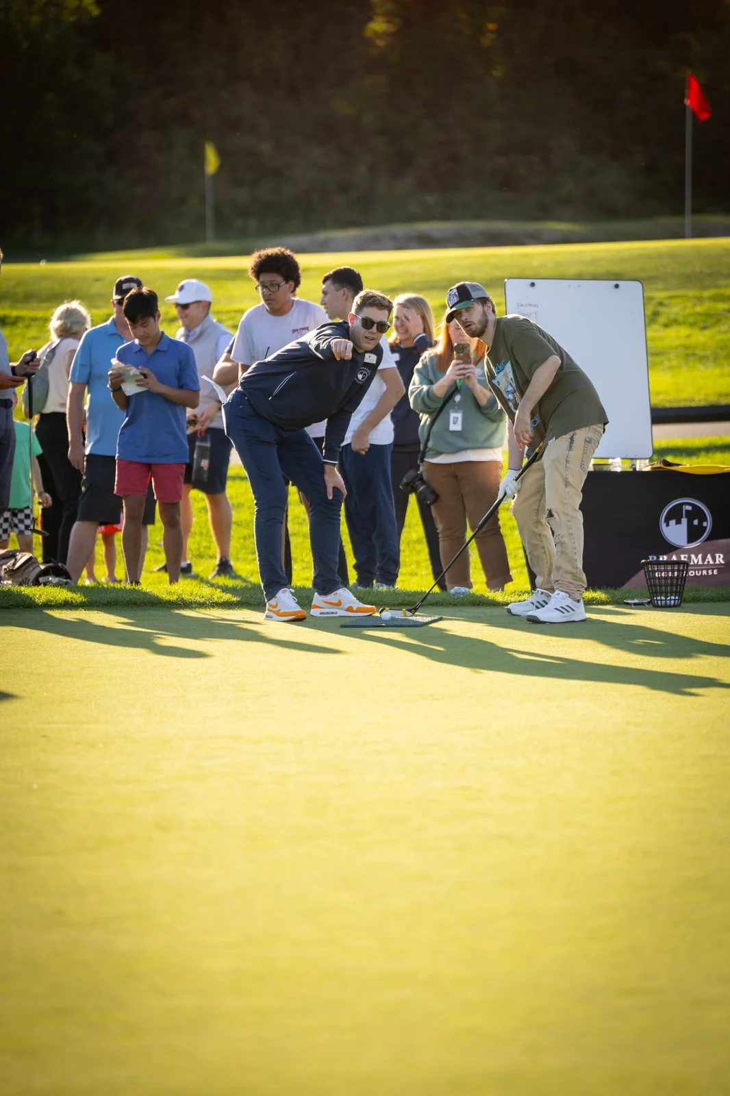 A group of people on a golf putting green. One man points to a golf ball while another with a putter watches, as others ob...
