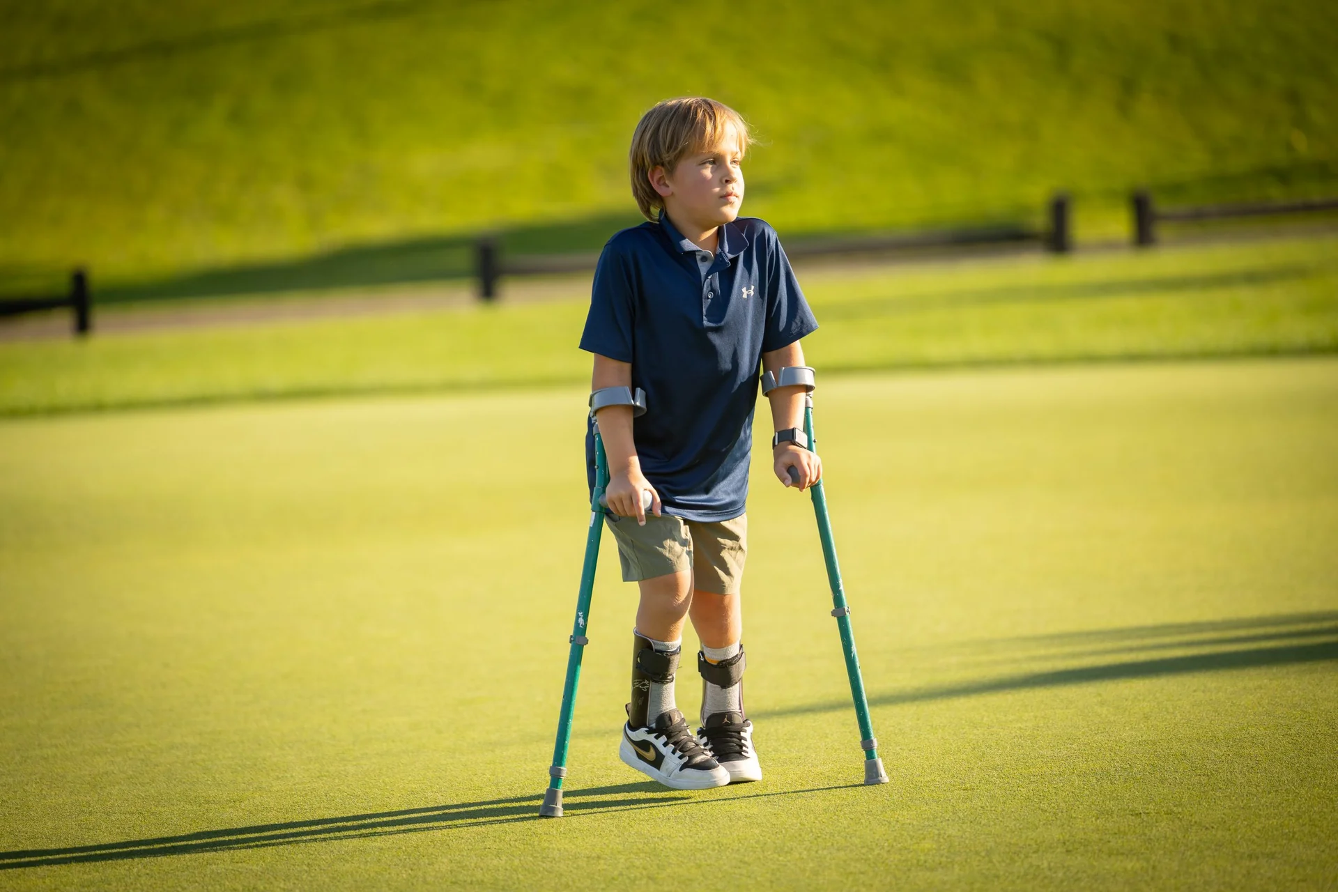 Young boy with leg braces and forearm crutches stands on a golf course green, looking right.