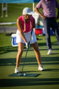 Woman in pink shirt and white skirt putting golf ball on a green practice mat.