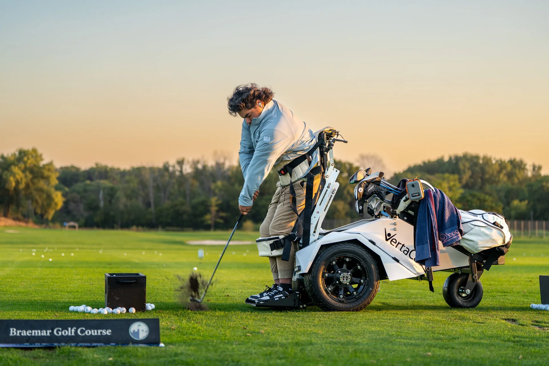 Golfer in adaptive stand-up cart swings club, kicking up divot on Braemar Golf Course practice range.