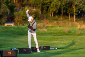 Golfer in follow-through swing on a sunny green course, with black bins bearing orange logos in the foreground.