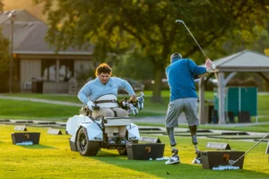Two golfers on a sunny driving range, one in an adaptive cart, the other with prosthetic legs swinging a club.