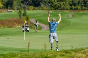 Golfer with prosthetic legs jubilantly celebrates on the green, raising arms and club. Other golfers and cart path in back...