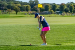 Female golfer with a prosthetic arm on a green fairway, about to hit a pink golf ball.