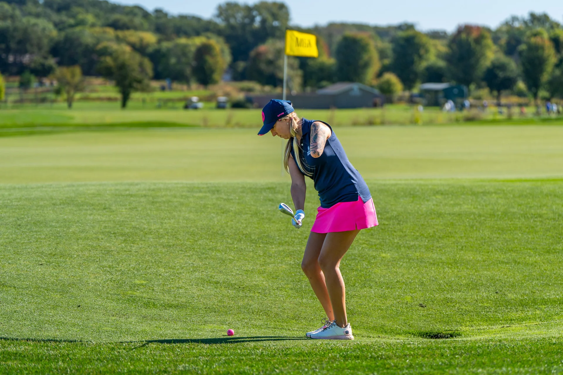 Female golfer with a prosthetic arm on a green fairway, about to hit a pink golf ball.