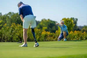 Golfer with prosthetic leg putts on a green as another golfer crouches, watching his line.