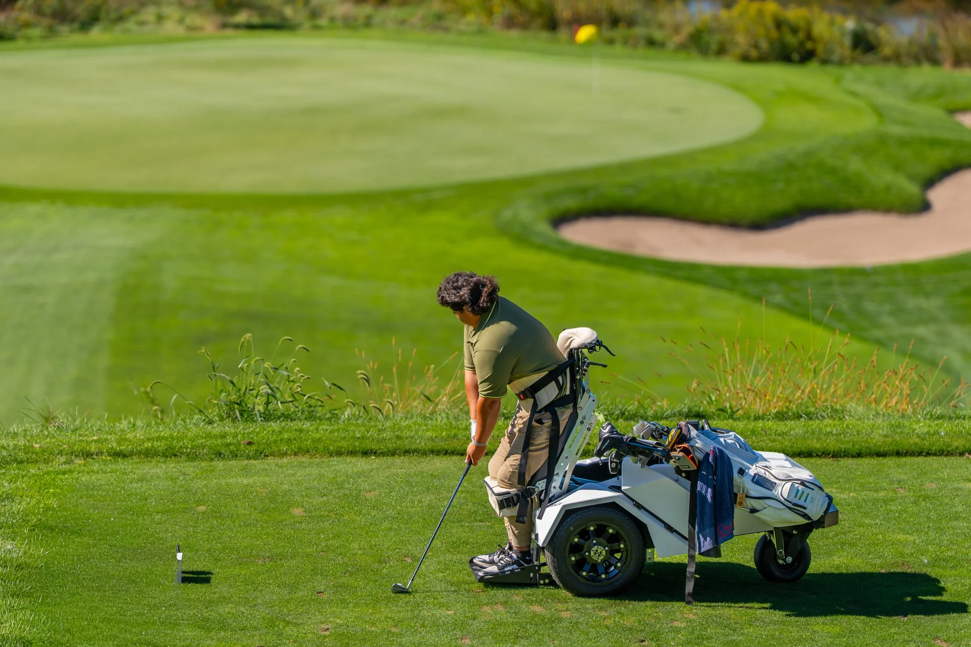 Golfer in adaptive standing cart prepares to hit on a sunny course, green and bunker in background.