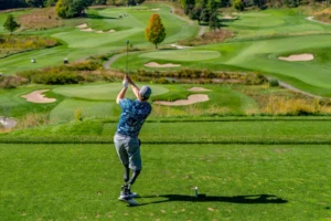 Golfer with prosthetic leg swings driver on tee box overlooking a vast, hilly golf course with multiple fairways and bunkers.