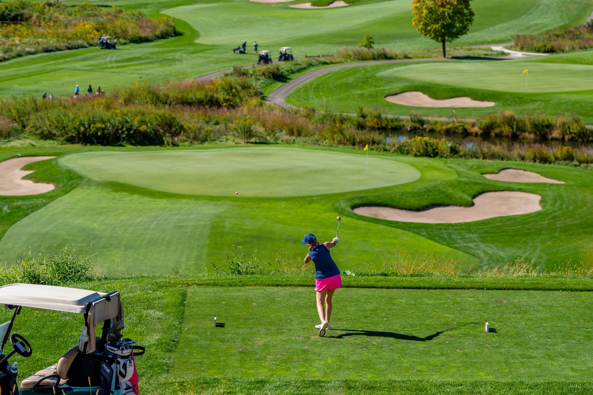 Golfer drives from a tee box towards a green with bunkers and a water hazard, overlooking a tiered golf course.