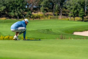 Golfer with prosthetic leg crouches on green, lining up a putt on a sunny course.