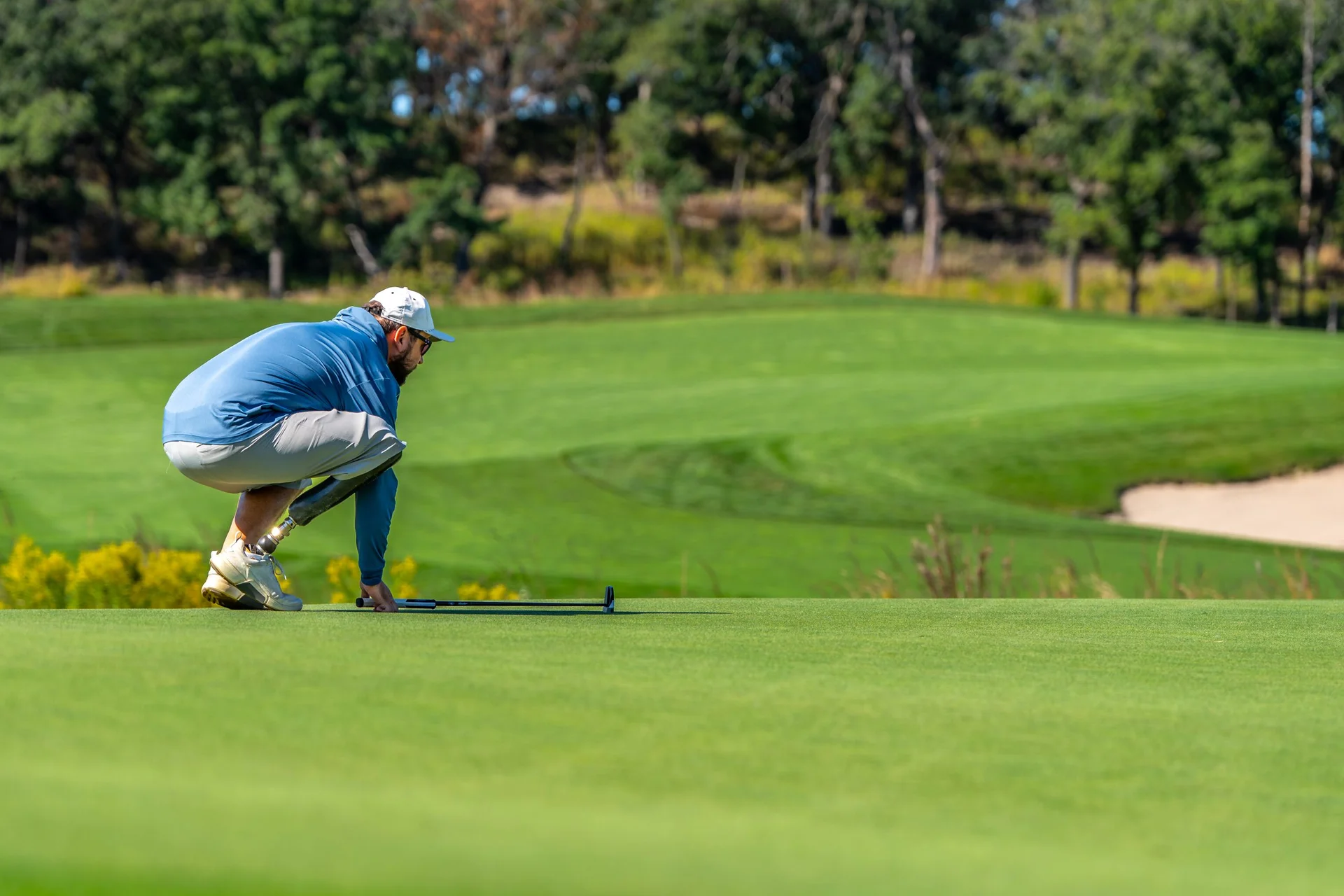 Golfer with prosthetic leg crouches on green, lining up a putt on a sunny course.