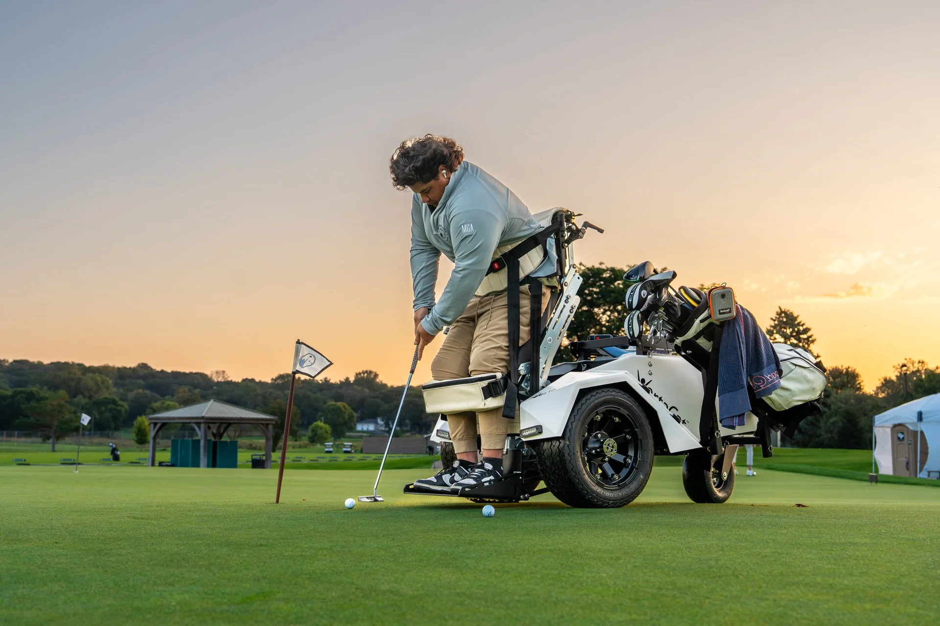 Golfer stands with adaptive equipment to putt on a golf course green during sunset.