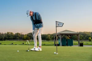 A golfer in a cap and blue shirt practices putting on a green, with a golf flag and driving range facilities.