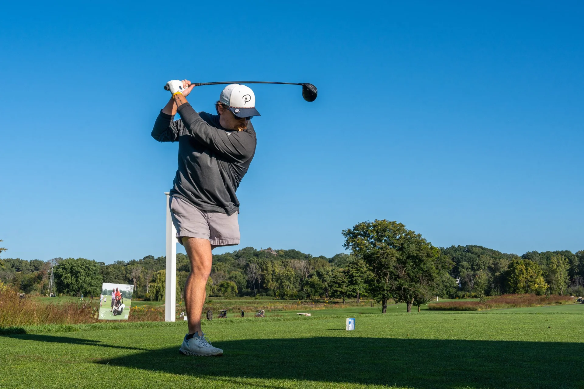 Golfer in grey shirt and white hat mid-swing with a driver on a sunny green golf course.