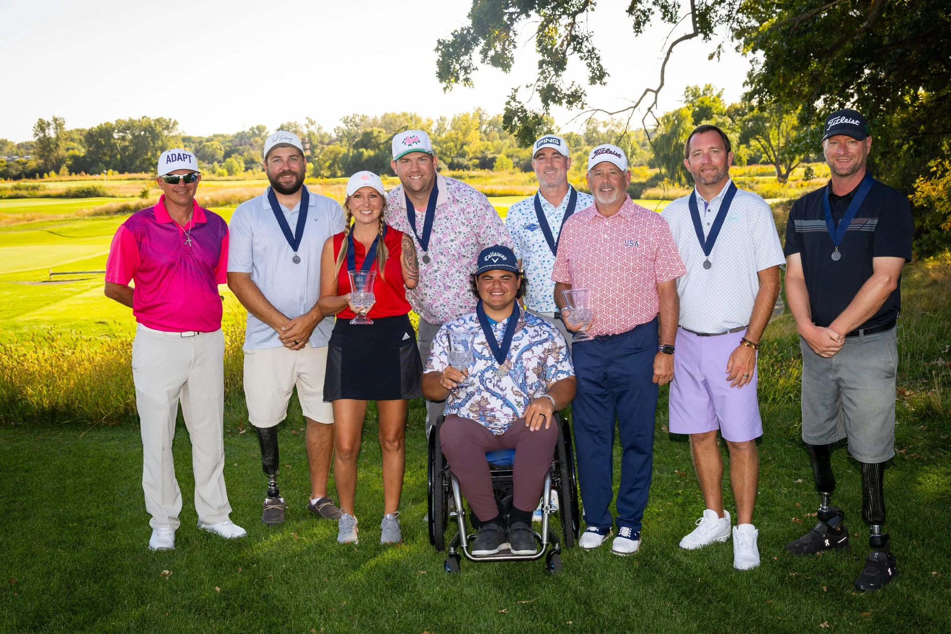 Nine adaptive golfers, some with prosthetics or in a wheelchair, stand on a course with medals and trophies, smiling.