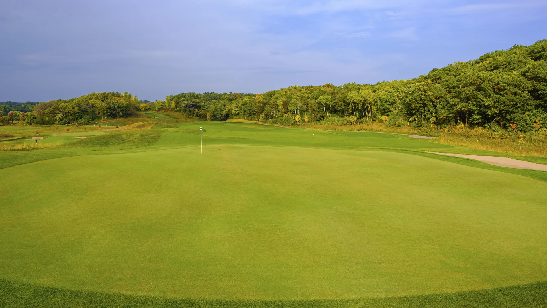 Green putting surface with a flagstick, fairway leading to tree-covered hills under a partly cloudy sky.