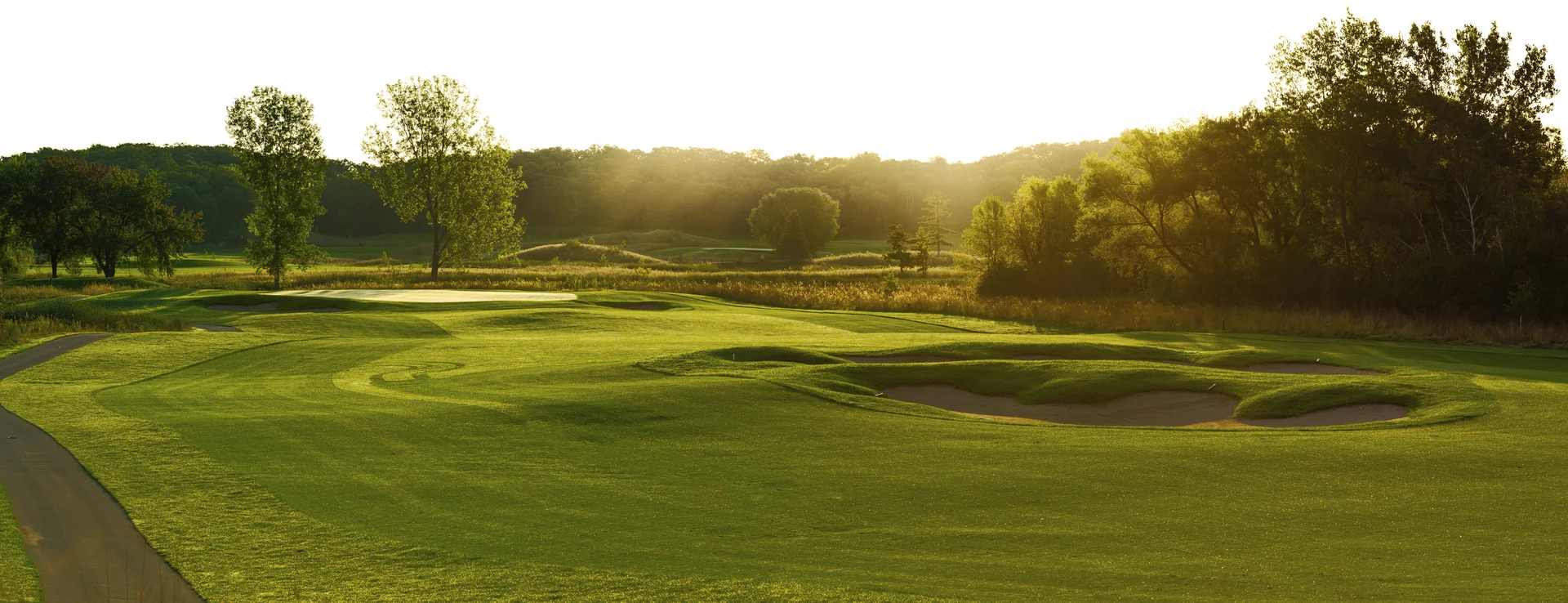 Sunny golf course with green fairways, sand bunkers, a cart path, and distant trees under golden light.