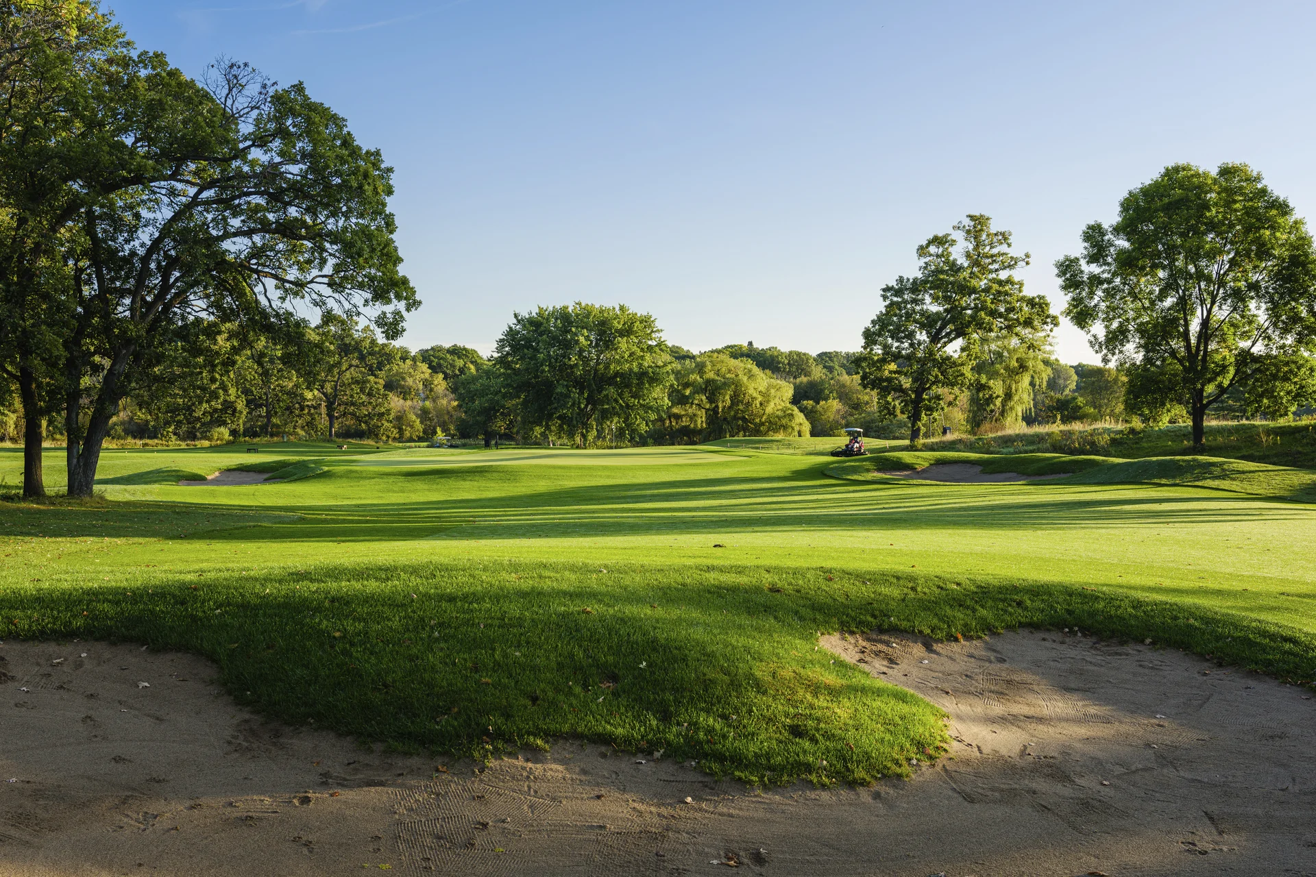 Golf course fairway with two prominent sand bunkers, framed by tall trees under a clear blue sky. A golf cart is visible i...