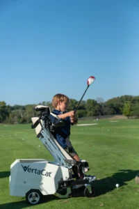Child in VertaCat adaptive golf chair swings a driver on a sunny golf course driving range.