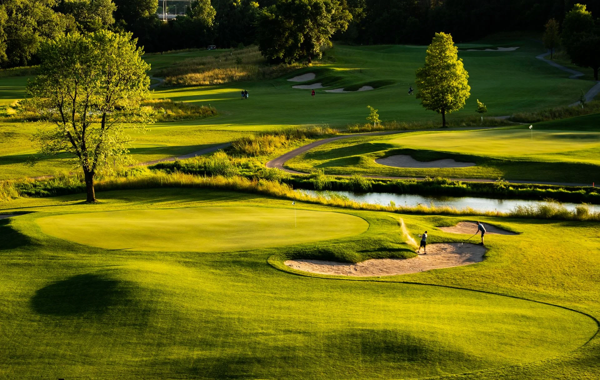Aerial view of a sunny golf course with a green, bunker, pond, and golfers playing on a golden lit fairway.