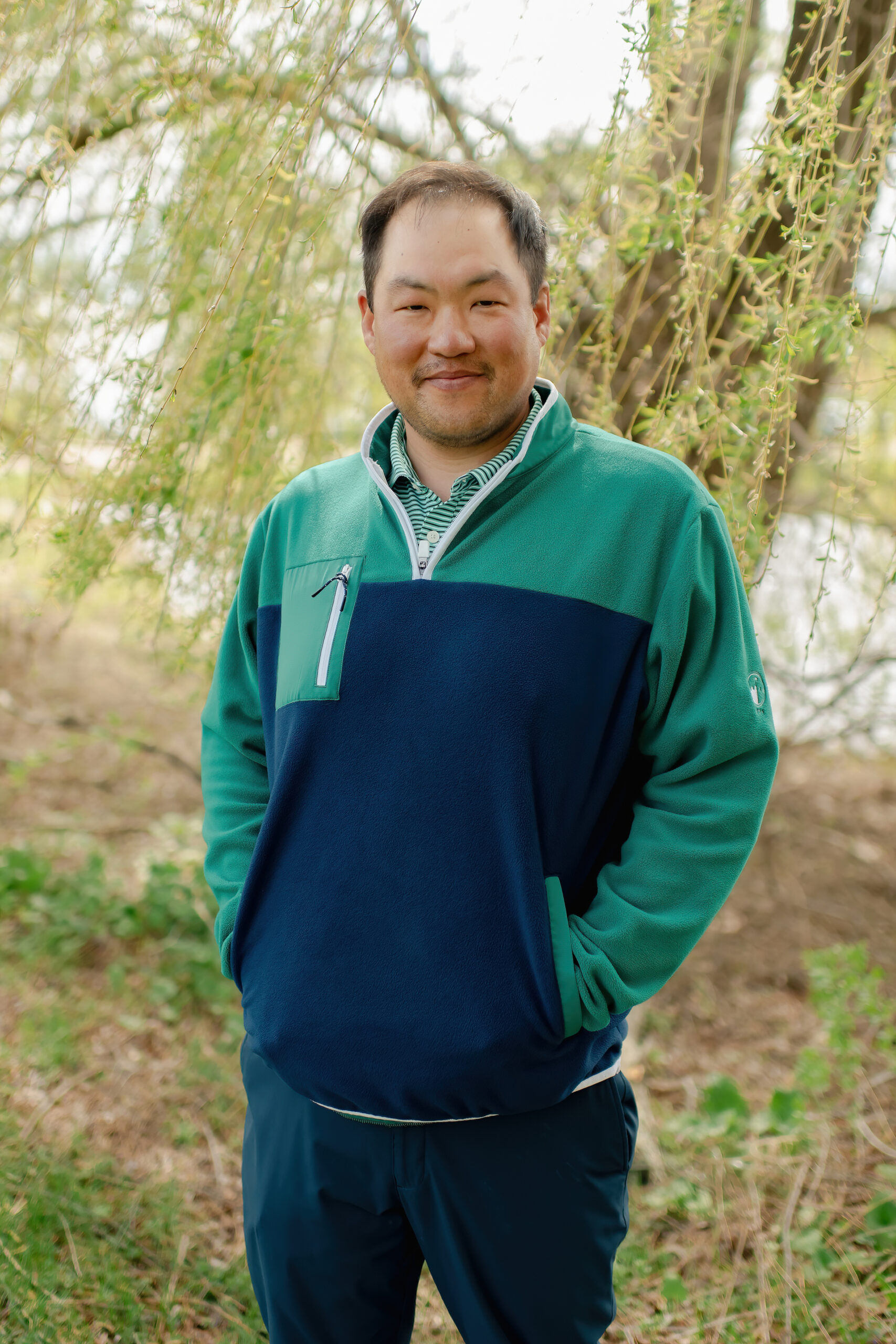 A smiling man with short dark hair standing in an outdoor natural setting. He is wearing a navy blue and green quarter-zip pullover over a green striped collared shirt.