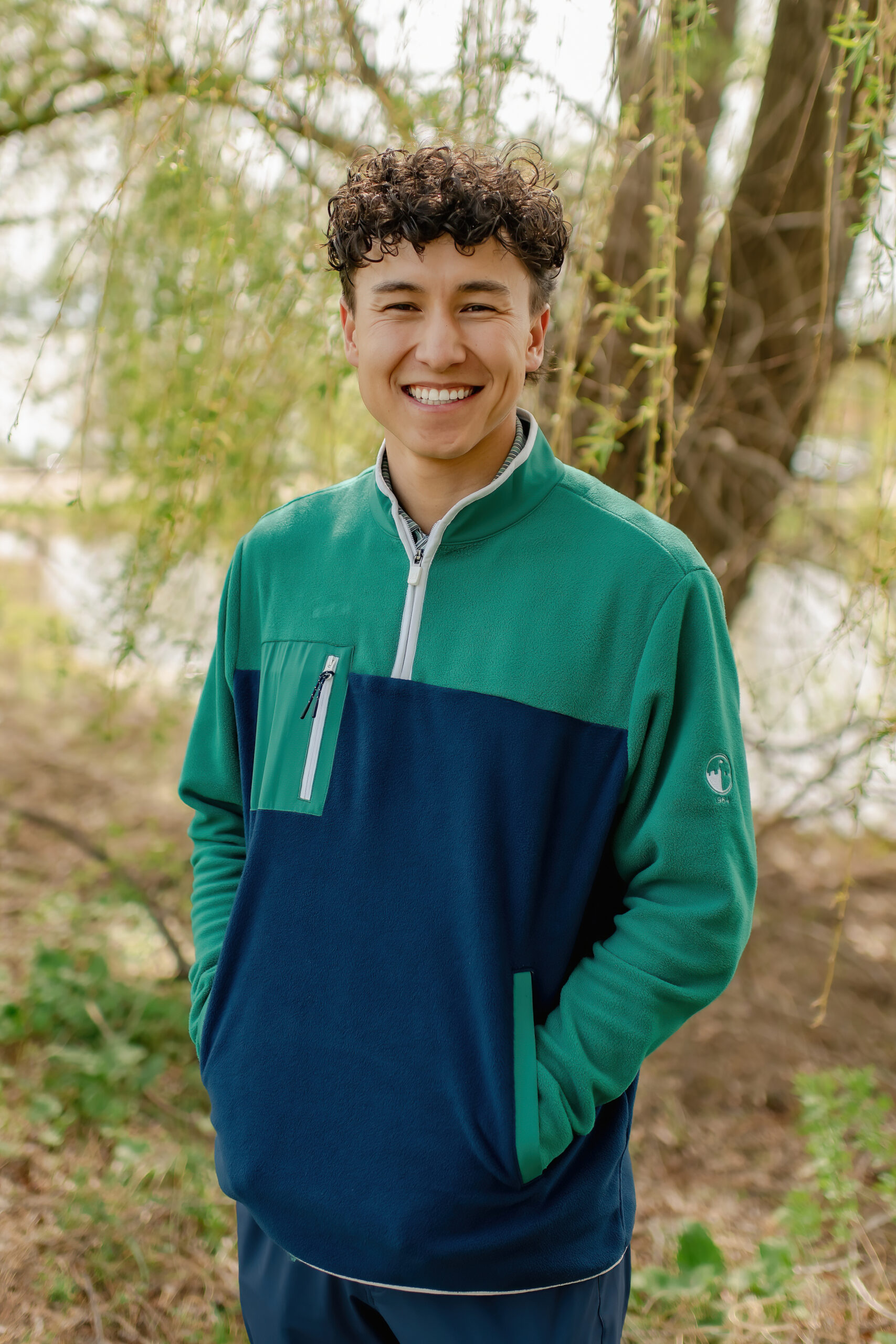 A smiling young man with dark, curly hair standing outdoors. He is wearing a signature navy and green quarter-zip fleece pullover with his hands in his pockets.
