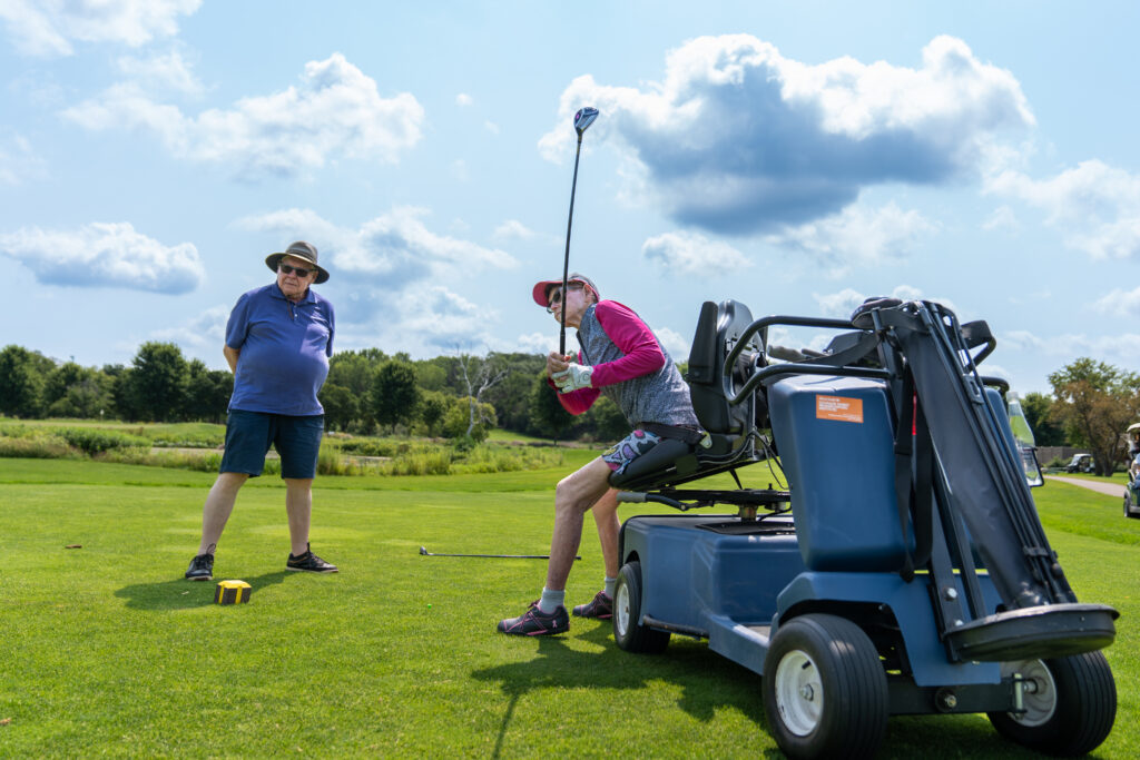 A golfer seated on a blue adaptive golf cart prepares to tee off on a sunny day. The cart’s seat is elevated and swiveled toward the ball, providing a stable platform for the swing. Another golfer stands nearby watching the shot.