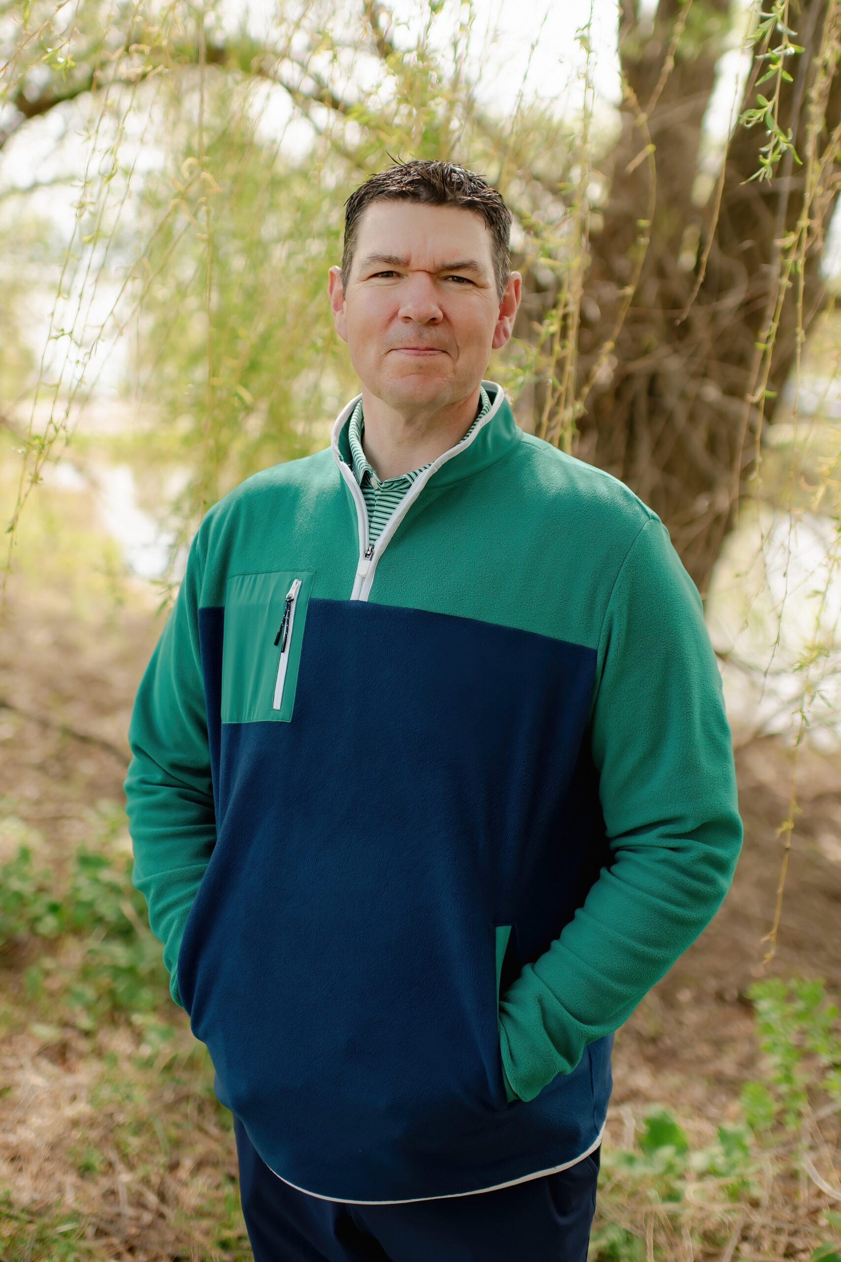 A man with short dark hair looking directly at the camera with a neutral expression. He is standing outdoors near a body of water, wearing a navy and green quarter-zip fleece.