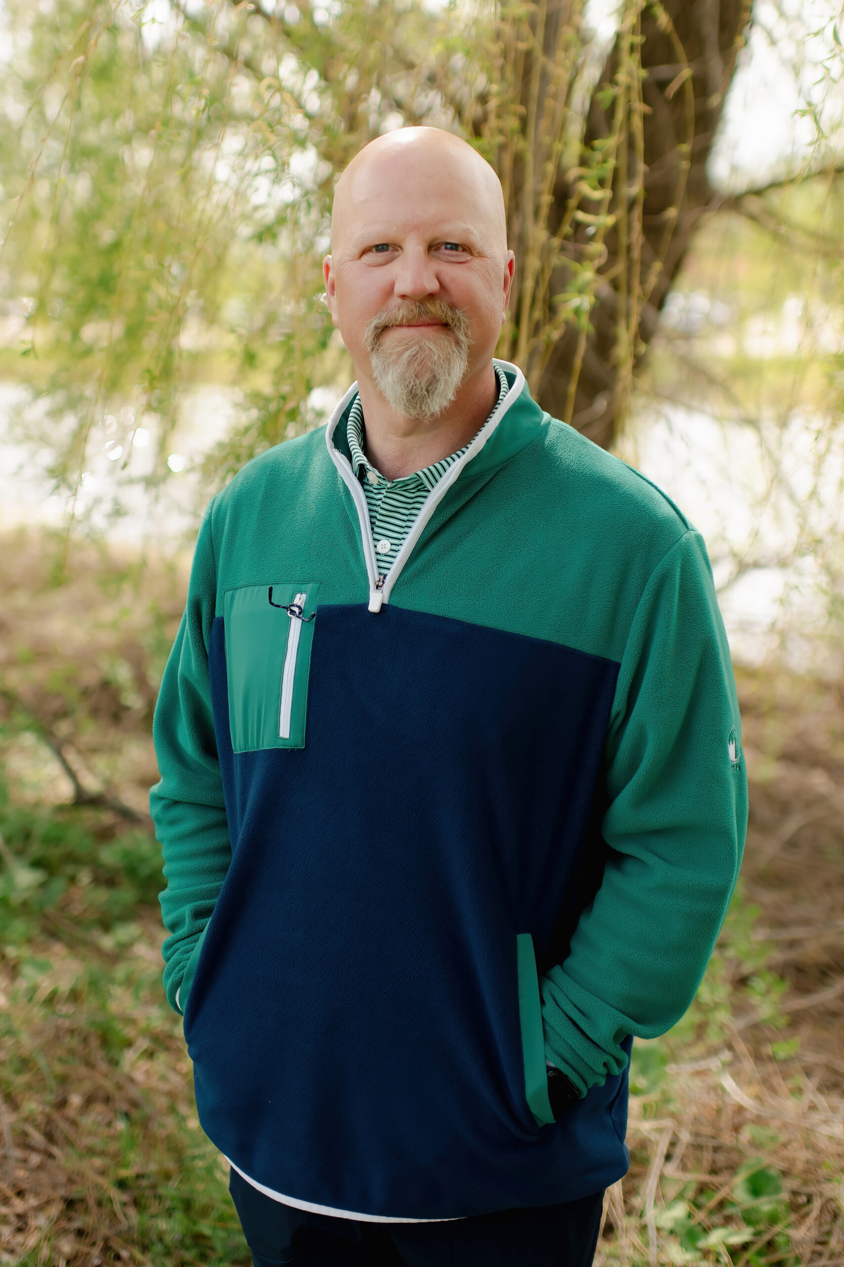 A man with a shaved head and a blonde goatee smiling for a portrait outdoors. He is dressed in a navy blue and green quarter-zip pullover, standing in front of a weeping willow tree.