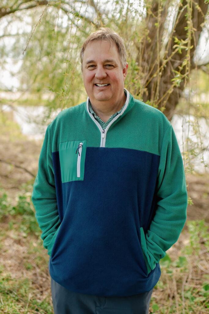 A smiling man with short brown hair standing in a natural outdoor setting. He is wearing a navy and green quarter-zip fleece jacket with a small zippered chest pocket.