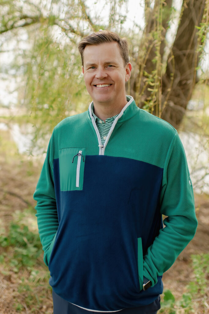A man with short brown hair and a friendly smile standing outdoors against a soft-focus background of trees and water. He wears a two-tone green and navy blue quarter-zip pullover.