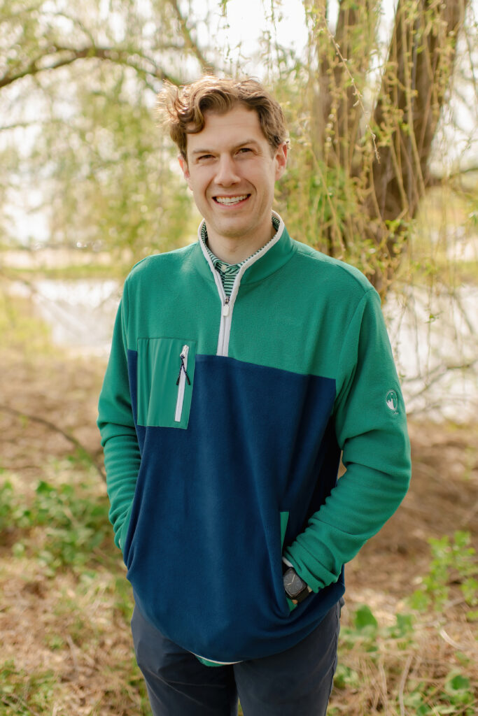 A smiling man with wavy brown hair outdoors, wearing a green and navy blue quarter-zip fleece over a striped collared shirt. He is standing in front of a willow tree with his hands in his pockets.