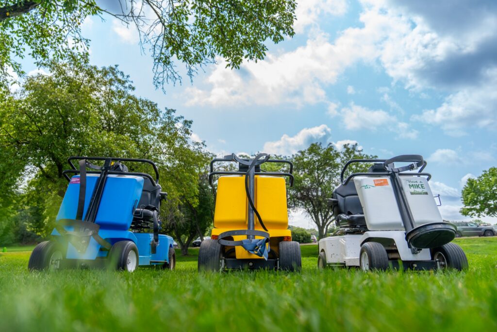 A low-angle row of three VertaCat adaptive golf carts—blue, yellow, and white—parked on a grass field. The carts feature specialized seating, harnesses, and standing mechanisms designed to help golfers with mobility impairments.
