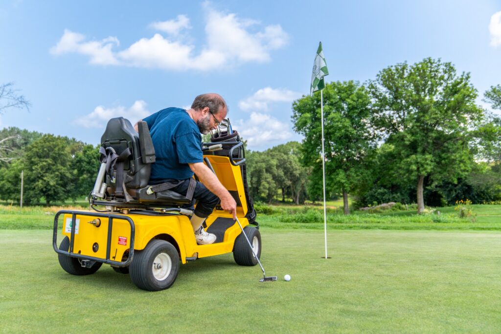A man using a yellow VertaCat adaptive golf cart on a green. He is seated in a specialized chair that has swiveled to the side, allowing him to lean over and use a putter to hit a golf ball toward a hole with a checkered flag.