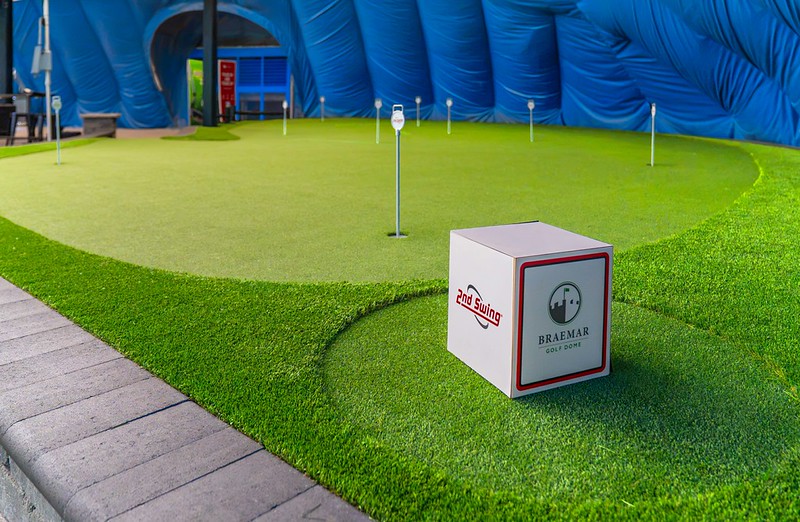 Wide view of an indoor artificial turf putting green with multiple practice holes and a branded 2nd Swing and Braemar Golf Dome block sitting in the foreground.