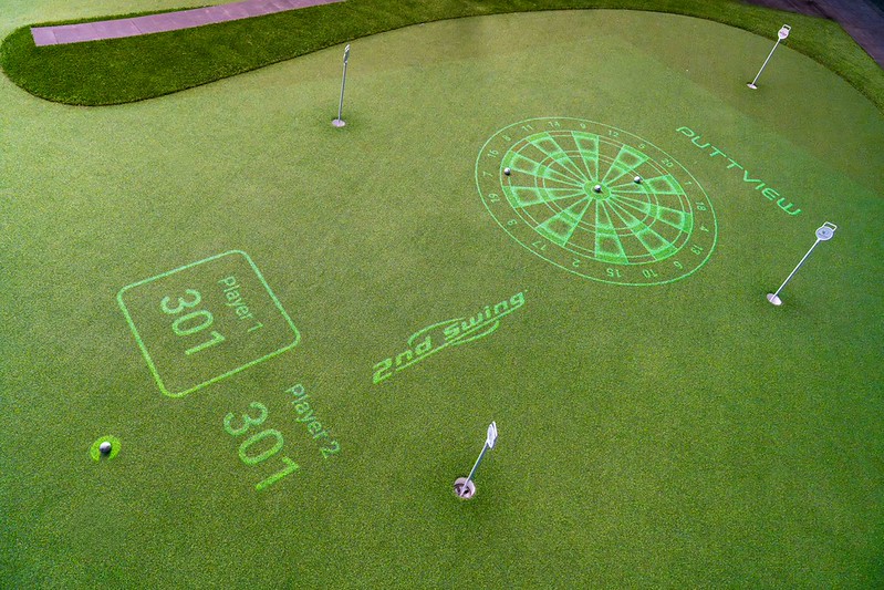 Overhead view of an indoor artificial turf putting green featuring a projected green dartboard game and player score displays.