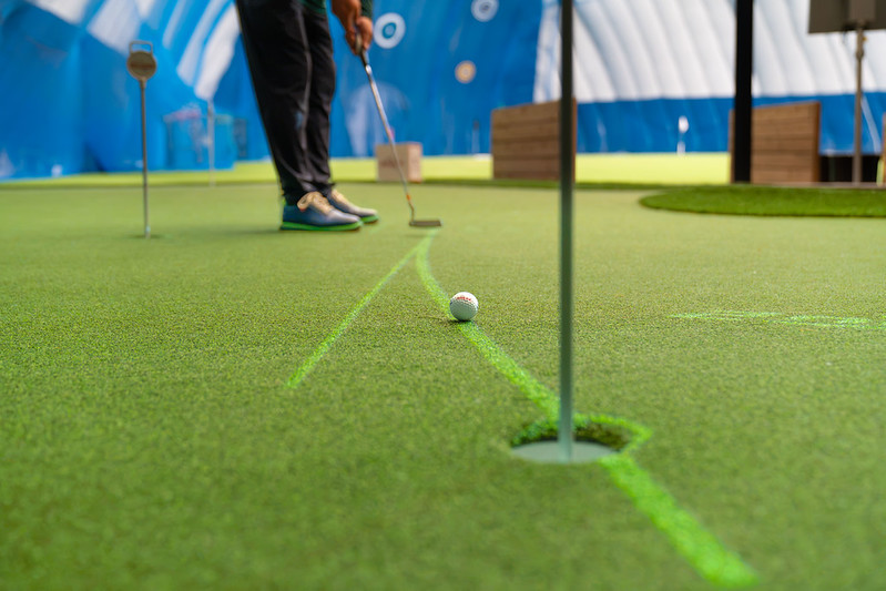 Low-angle shot of a golf ball rolling along a projected curved green putting line toward a hole on an indoor turf green, with a golfer in the background.
